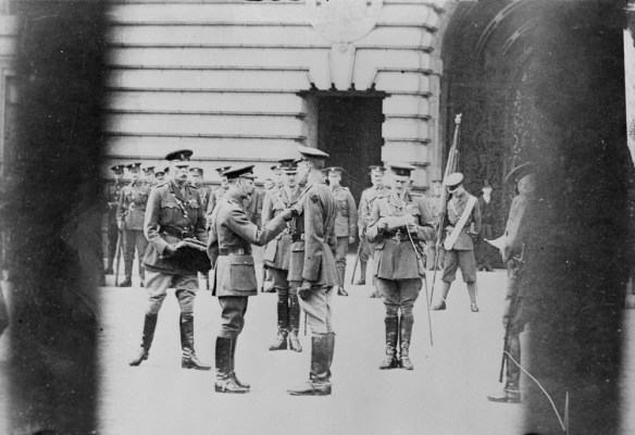 Photographie en noir et blanc d’un homme épinglant une décoration sur la poche de la veste d’un autre homme. Un autre homme lit la citation, alors qu’un quatrième tient une mallette. À l’arrière-plan, des soldats se tiennent au repos.