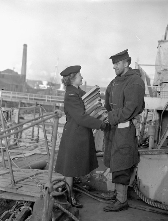 Photo en noir et blanc d’une jeune femme en uniforme donnant une grande pile de livres à un marin. Ils sont debout sur le pont d’un bateau et le havre figure en arrière-plan.