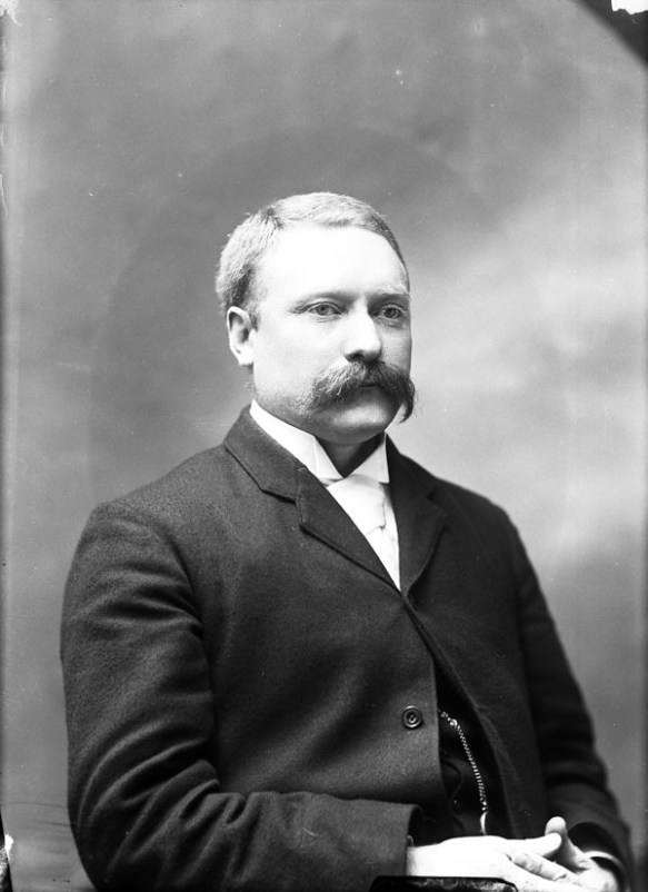 Photo noir et blanc montrant un homme moustachu portant un complet et une chemise blanche.