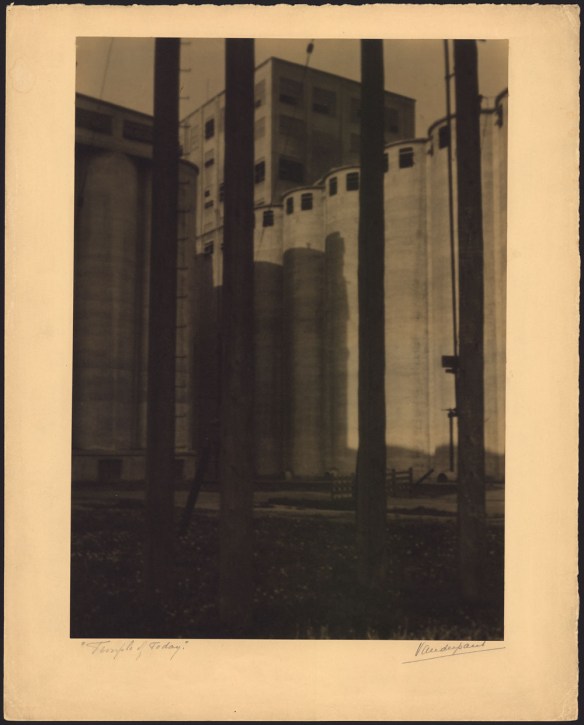 Photographie noir et blanc d’un silo à grains; de hautes tours circulaires se dressent devant un immeuble rectangulaire encore plus haut.