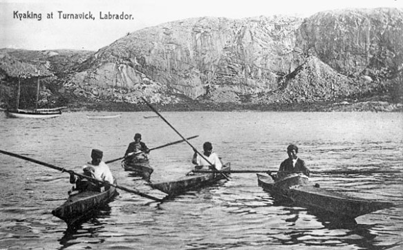Photographie noir et blanc montrant quatres hommes dans des kayaks avec un grand rocher au lointain.