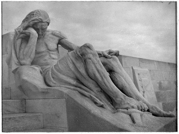 Photographie noir et blanc d’une imposante sculpture du Mémorial de Vimy, représentant un homme endeuillé dont le pied repose sur une épée. En arrière-plan, on voit des panneaux où sont inscrits les noms des Canadiens morts au champ d’honneur.