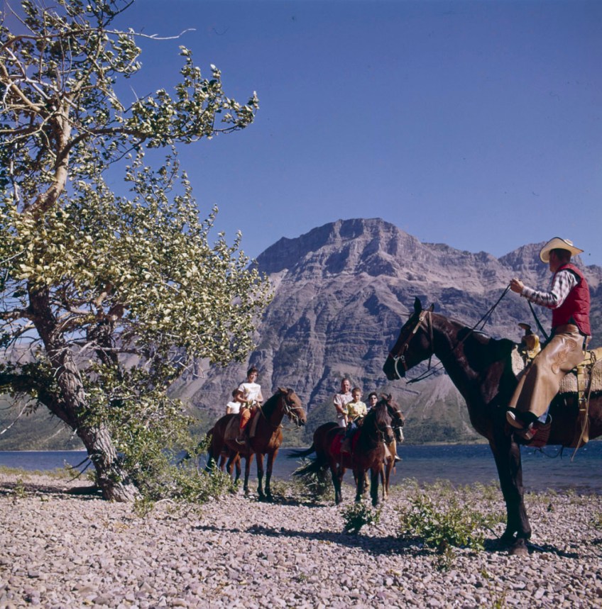 Photographie couleur d’un groupe de personnes à cheval, près d’une rivière. L’un des cavaliers, habillé en cow-boy, semble guider une famille sur un sentier. On voit un pic montagneux en arrière-plan.