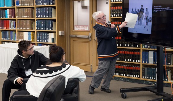 Photographie en couleurs d’un aîné inuit qui porte une veste traditionnelle inuite, se tient devant un moniteur et pointe une image à l’écran. Deux jeunes inuits regardent et écoutent attentivement.