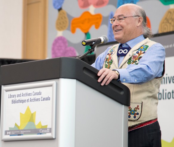 Photographie en couleurs d’un homme de la Nation métisse portant une veste ornée de broderies et se tenant sur un podium. 