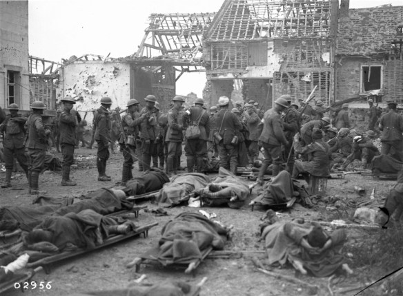 Photo noir et blanc montrant une scène de guerre : des soldats blessés sont étendus sur des civières; autour deux, plusieurs soldats se tiennent debout devant des bâtiments en ruines. 