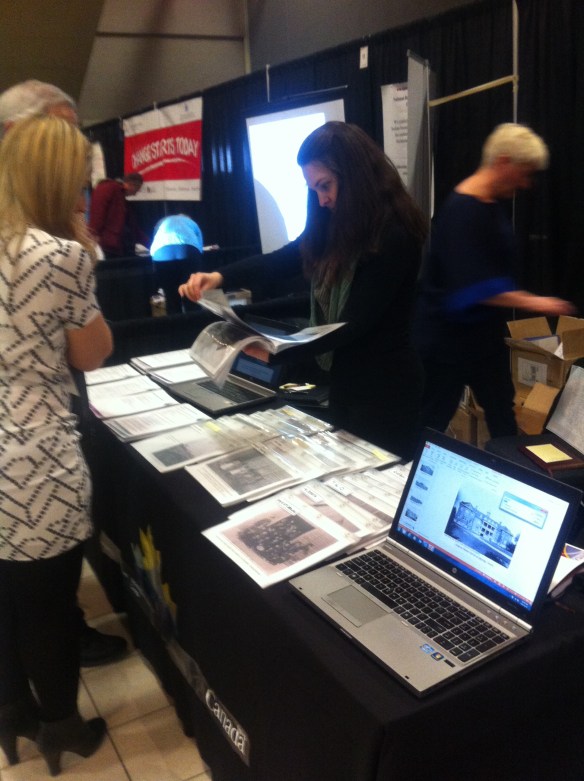 Photo d’un kiosque présentant de nombreuses photos et un ordinateur sur le bord d’une table. Une dame aux cheveux bruns qui travaille au kiosque trouve une photo pour un couple. Un autre kiosque est visible à l’arrière-plan.