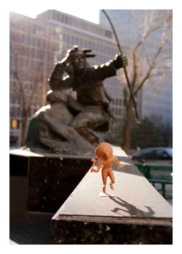 Petite figurine marron clair d’un membre des Premières Nations placée devant la statue d’un chasseur indien. En arrière-plan, on peut voir de grands immeubles à bureaux et des arbres.