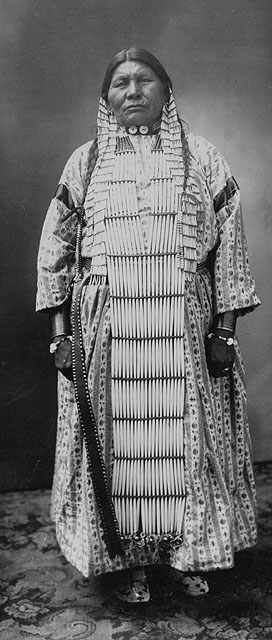 Photo en noir et blanc d’une femme de la Première Nation des Dakota portant une robe rayée sous un long plastron à perles. Elle porte également des tresses, un collier et de longues boucles d’oreilles. 