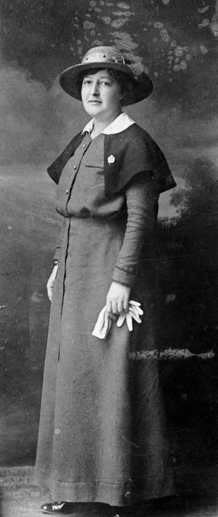 Une photo noir et blanc d’une femme dans l’uniforme d’une sœur infirmière avec la cape, l’épinglette, le chapeau et les gants blanc. 
