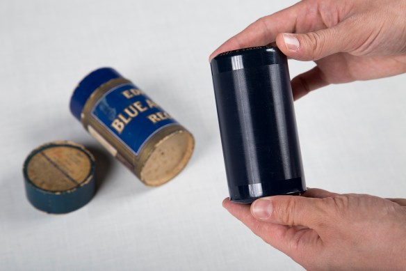 Une photographie couleur de mains tenant un cylindre bleu foncé couvert de sillons. La boîte en carton du cylindre se trouve à l’arrière-plan.