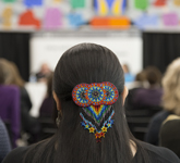 Photo couleur d’une pince à cheveux ornée de perles multicolores à l’arrière de la tête d’une femme. Celle-ci prend place dans la salle Pellan de Bibliothèque et Archives Canada, où elle écoute un groupe de conférenciers.