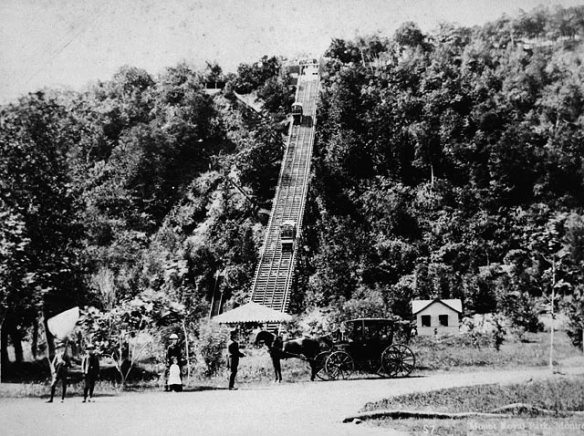 Photographie en noir et blanc d’un funiculaire montant une côte très boisée, en bas de laquelle se trouvent une voiture tirée par un cheval et quelques personnes regardant le photographe.