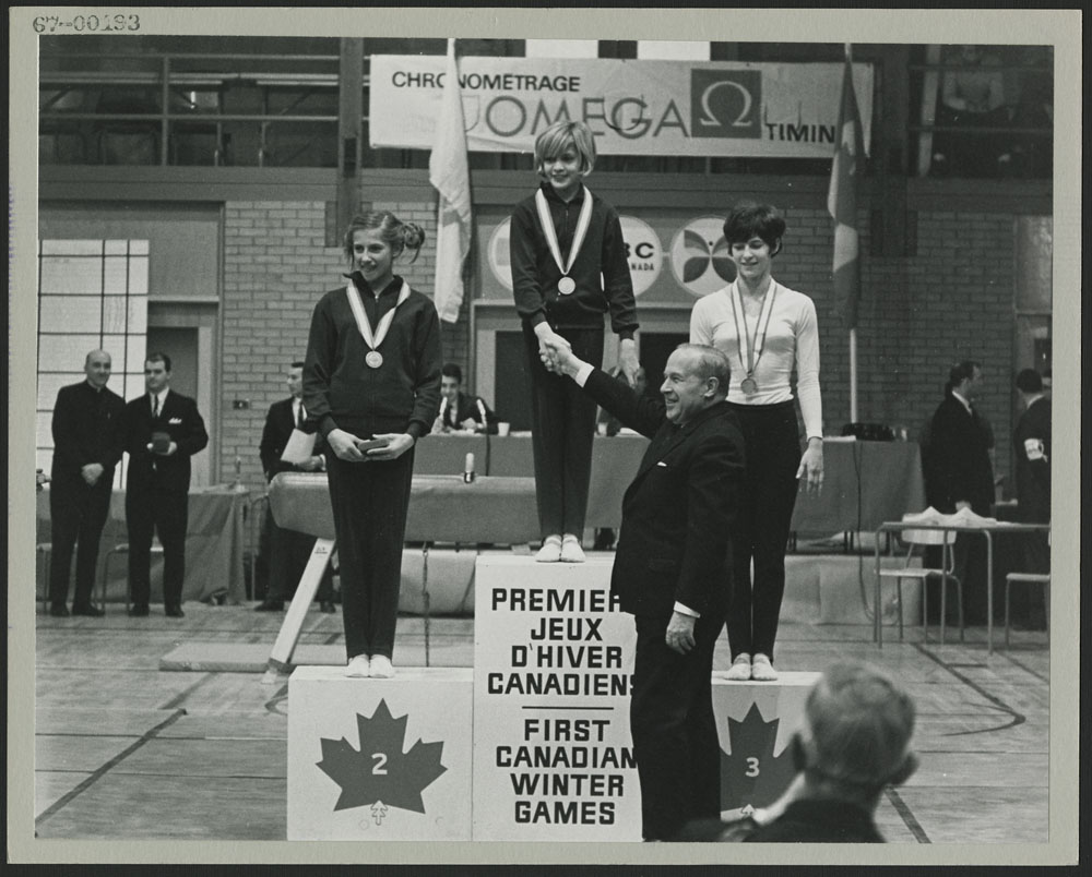 Photo noir et blanc montrant un podium avec trois jeunes femmes portant des médailles. Un homme serre la main de la médaillée d’or.