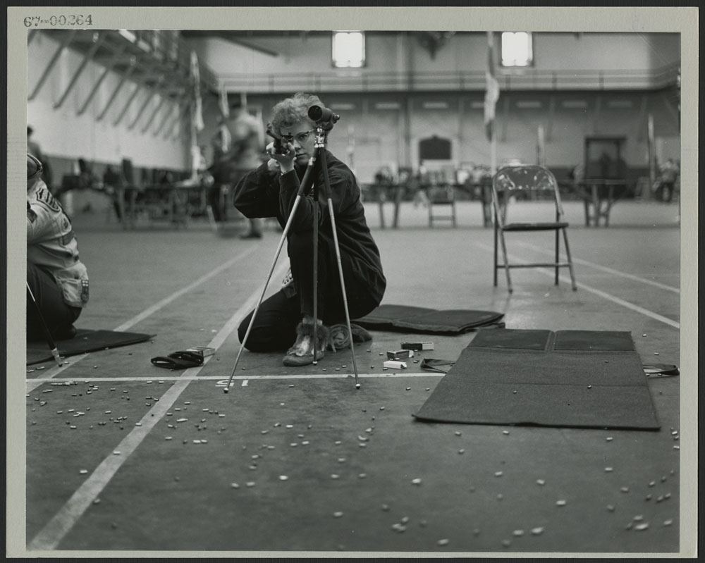 Une photo noir et blanc montrant une femme agenouillée visant avec une carabine; des cartouches vides sont parsemées sur le sol autour d’elle.