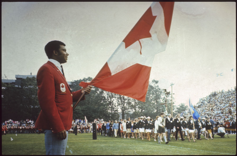 Photo en couleur d’un homme portant un veston rouge et tenant un étendard du drapeau canadien pendant que des gens paradent à l’arrière-plan.