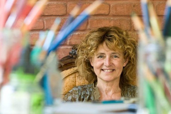 Photo en couleur d’une femme assise et souriante. On aperçoit dans l’avant plan des crayons de couleurs flou.