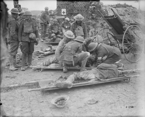 Photographie en noir et blanc de deux hommes étendus sur des brancards. Du personnel médical s’occupe d’un des deux blessés, alors que l’autre est étendu sur le côté. Plusieurs soldats se tiennent debout à gauche des brancards, et d’autres sont assis à l’arrière-plan. La scène se passe dans les décombres d’un édifice bombardé dont il ne reste qu’une cheminée.