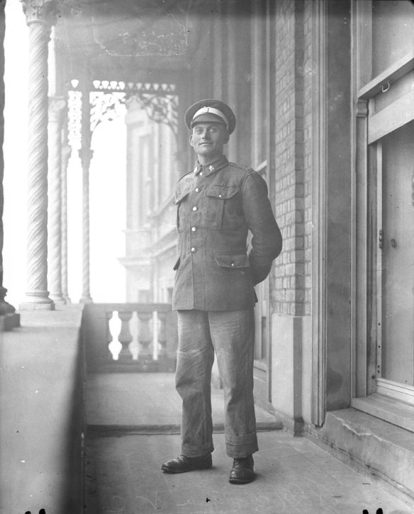 Photographie en noir et blanc d’un jeune homme en uniforme, debout sur un balcon à l’extérieur, les mains derrière le dos. 