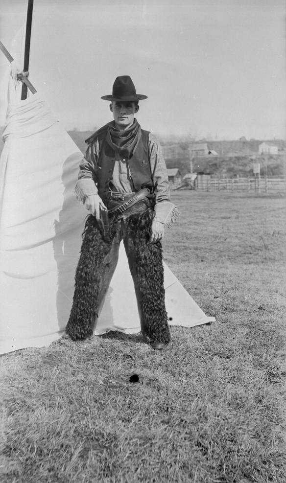 Un photo en noir et blanc d’un cowboy portant un chapeau noir, un bandana, des gants et des jambières de fourrure. Il est debout devant une tente, la main droite posée sur un pistolet dans son étui.