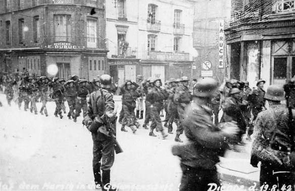 Photo noir et blanc de soldats canadiens capturés, marchant en rang dans une ville sous la surveillance de soldats allemands.