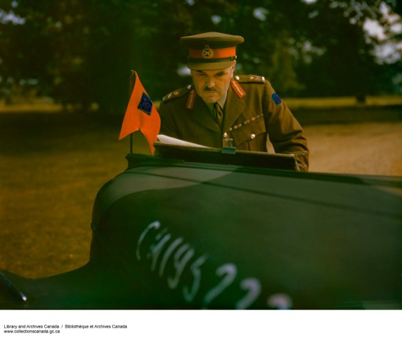 Photo couleur du major général J. H. Roberts en uniforme, examinant des documents sur le capot d’une voiture d’état-major.