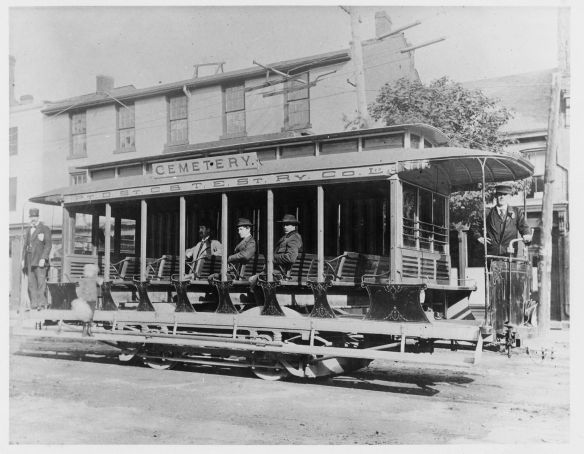 Une photographie en noir et blanc d’un tramway à ciel ouvert. Il y a un conducteur à l’avant et à l’arrière du tramway et trois passagers sont assis. Un garçon est debout sur la banquette latérale.