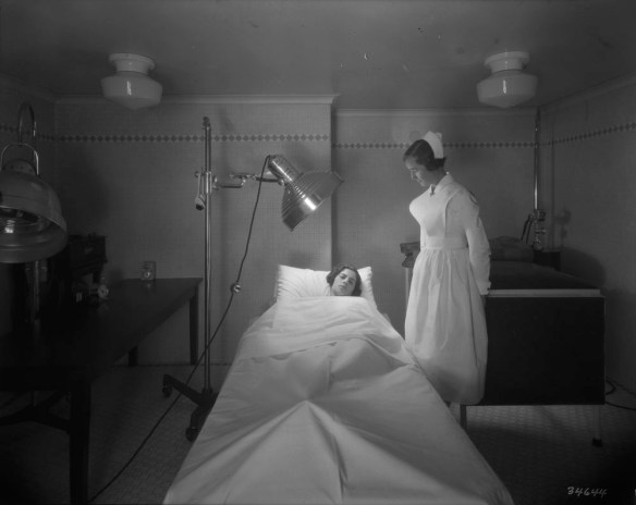 Une photographie en noir et blanc d’une infirmière prenant soin d’une patiente qui reçoit un traitement d’une lampe infrarouge. La patiente est allongée sur un lit.