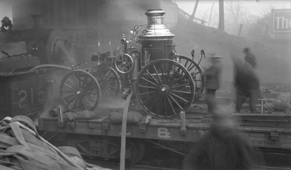 Photographie en noir et blanc d’une pompe à incendie à vapeur sur un wagon plat. Des hommes utilisent la pompe pour combattre un incendie près d’une voie ferrée et de remises.