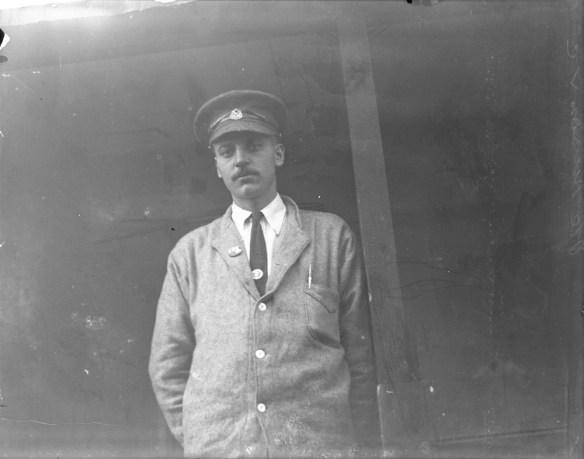 Une photographie en noir et blanc d’un homme portant une casquette, une veste de laine grise, une chemise blanche et une cravate foncée.