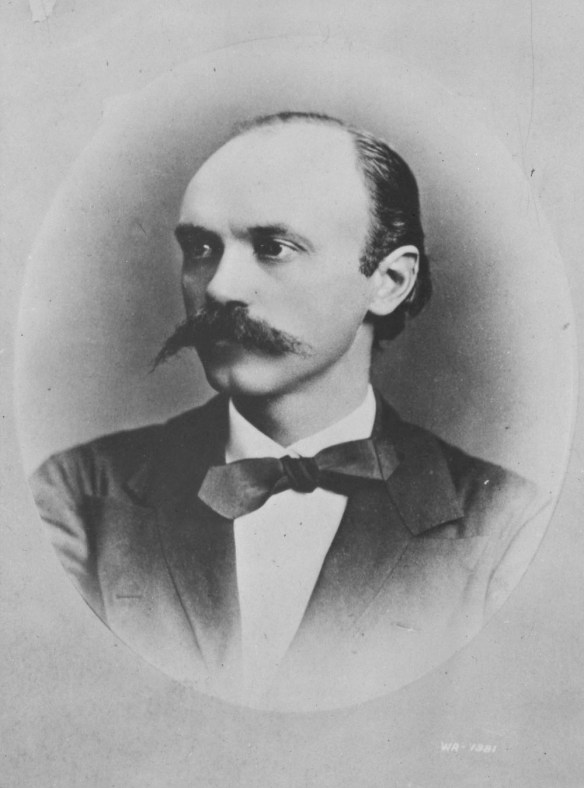 Photo noir et blanc d’un homme à la moustache proéminente portant un veston et un nœud papillon. La photo de forme ovale est placée sur un carton de montage mat de couleur grise.