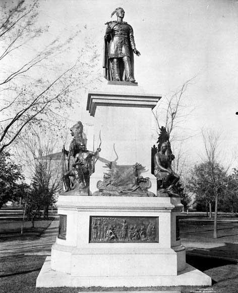 Une photo noir et blanc d’une statue de bronze sur un piédestal en pierre orné de reliefs et de statues de bronze sur tous les côtés. On peut voir à l’arrière-plan un parc aux arbres dénudés. 