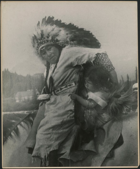 Femme à cheval, tenant une petite fille à ses côtés. La femme porte une tunique unie avec une ceinture à motif; ses cheveux tressés sont ornés d’une coiffe de plumes. 