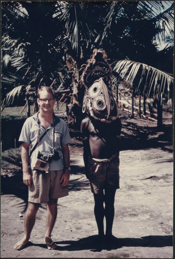 Photographie en couleurs d’un homme portant un short, une chemise et des sandales. Une caméra munie d’une courroie pend à son cou. Il se tient debout à côté d’un homme qui porte un short et un masque élaboré.
