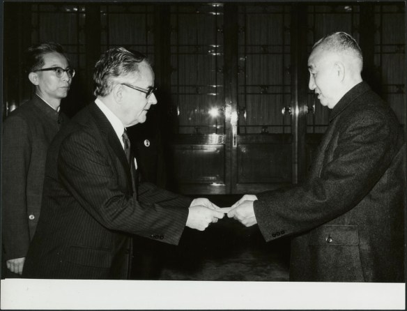 Photographie en noir et blanc de deux hommes qui se saluent. L’un des deux hommes tient un bout de papier et le tend à l’autre. Un homme se trouvant à l’arrière-plan regarde la scène.