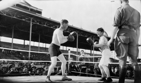 Photographie en noir et blanc de deux soldats qui s’affrontent à la boxe. L’un porte un short noir, et l’autre, un short blanc. Des soldats assis autour du ring assistent au combat.