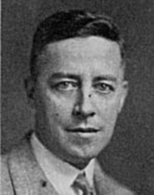 Photographie en noir et blanc d’un homme vêtu d’un veston gris qui fixe l’objectif. 