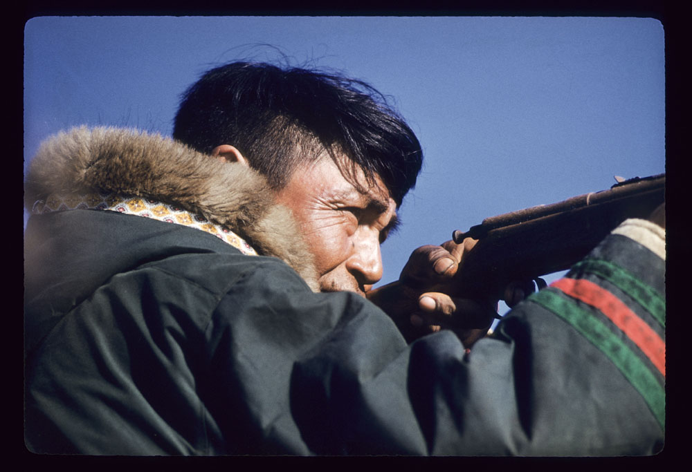 Photographie couleur d’un homme de profil tenant un fusil et ajustant son tir. Il porte un parka traditionnel au capuchon bordé de fourrure et aux manches ornées de galons verts et rouges.