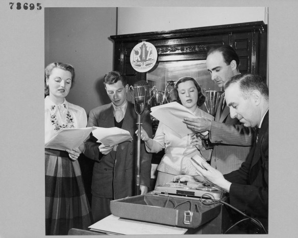 Photographie en noir et blanc de deux femmes et de trois hommes, membres de la troupe de radiothéâtre RA, lisant dans un microphone à partir d’un texte.