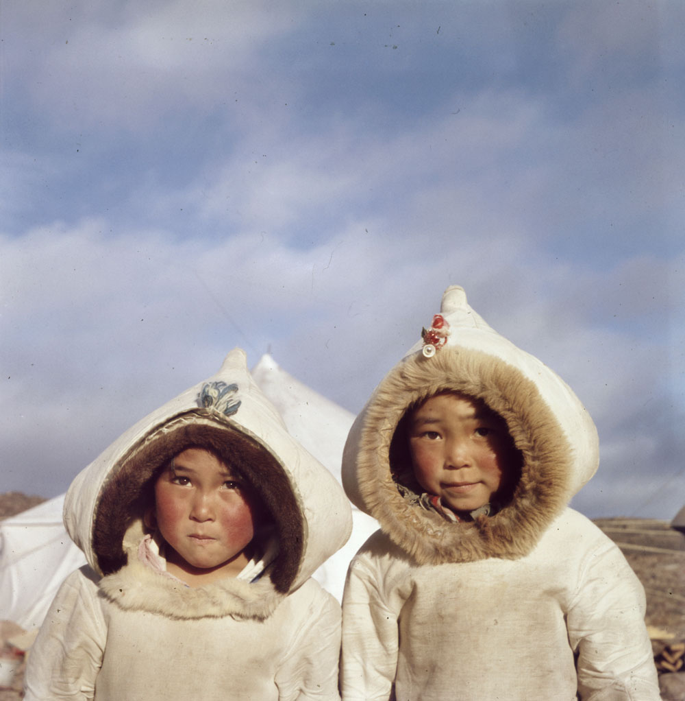 Photographie couleur de deux enfants inuits portant un manteau traditionnel devant une tente blanche dans un paysage rocailleux.