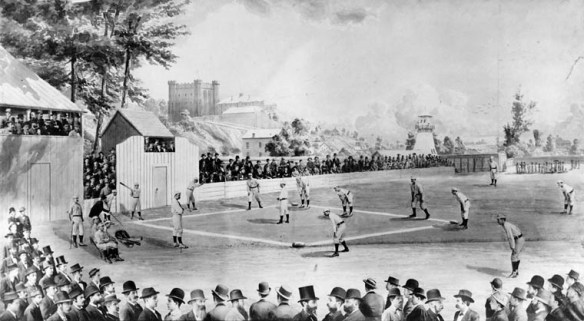 Une photographie noir et blanc d’un terrain de baseball extérieur où une partie est en cours. Une foule compacte y assiste dans les gradins. Des édifices de la ville se dessinent à l’arrière-plan.