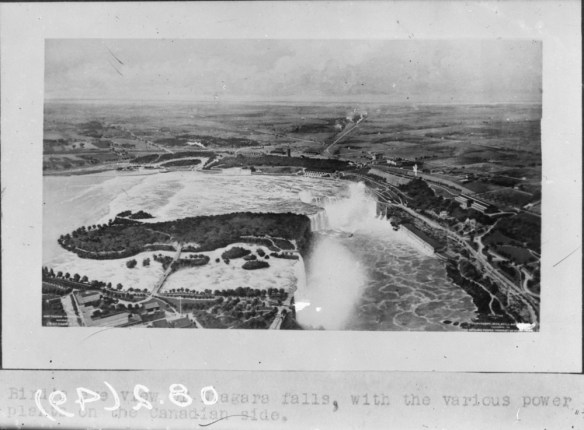 Une photographie en noir et blanc des chutes Niagara, à partir d’une vue à vol d’oiseau. On aperçoit divers bâtiments des deux côtés de la frontière ainsi que des routes menant aux rives. 
