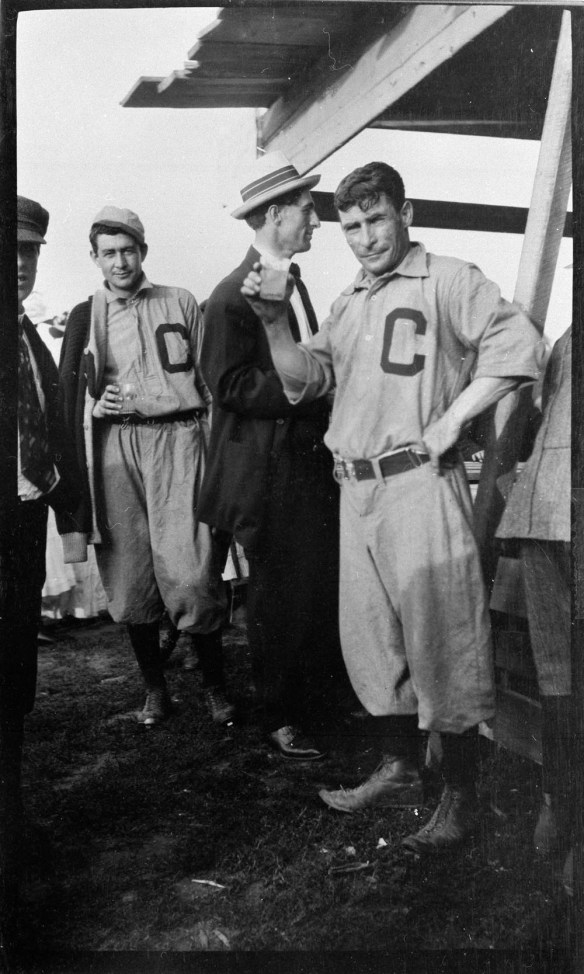 Photo noir et blanc de cinq hommes debout devant un comptoir. Deux d’entre eux sont vêtus d’un uniforme de baseball orné d’un grand « C » sur la poitrine; les autres portent un complet et un chapeau. Un des hommes en uniforme tient une boisson dans sa main et regarde l’objectif.