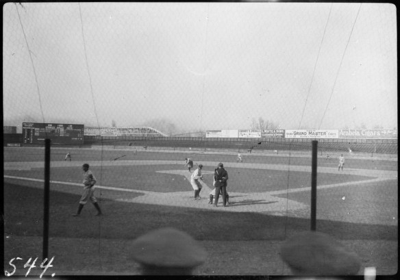 Une photographie noir et blanc d’une partie de baseball prise de l’arrière du marbre. Un joueur est au marbre, prêt à recevoir un lancer. L’arbitre est debout derrière le receveur pour rendre sa décision.