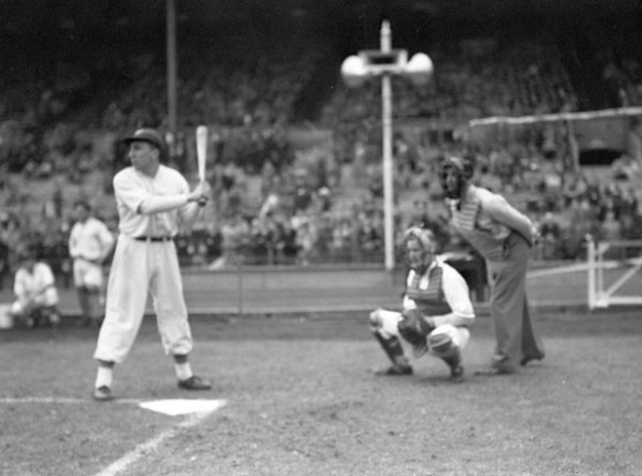 Une photographie noir et blanc d’une partie de baseball. Le frappeur est debout levant un bâton et, derrière lui, le receveur et un arbitre. À l’arrière-plan, on aperçoit des joueurs guettant le jeu et des spectateurs dans les gradins.