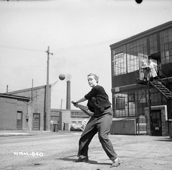 Une photographie noir et blanc d’une femme portant des vêtements de travail et un foulard noué sur la tête s’apprête à frapper une balle avec son bâton. Elle est debout dans la cour arrière d’un bâtiment industriel, et l’on voit d’autres structures à l’arrière-plan.