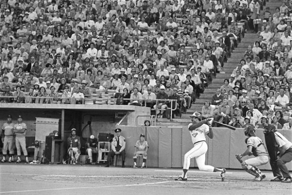 Photo noir et blanc d’une partie de baseball. Un homme vient de frapper la balle. Derrière lui, on aperçoit un receveur accroupi, dos à un arbitre, lui aussi accroupi. Des joueurs de baseball en uniforme et un policier observent la scène en retrait. Derrière eux, les gradins sont remplis de spectateurs.