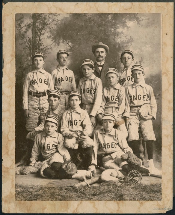 Une photographie noir et blanc de 10 enfants vêtus d’un uniforme de baseball. Les chandails portent l’inscription « Pages » sur le devant. Certains des garçons sont assis et d’autres debout avec des bâtons, des gants et d’autres équipements de baseball. Un homme adulte, portant un costume et un chapeau, se tient debout derrière les garçons. L’arrière-plan est une toile de fond montrant des arbres.