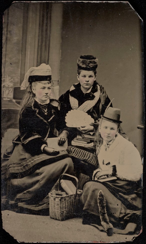 Photo noir et blanc de trois femmes prenant la pose pour un portrait. Elles sont vêtues de longues robes, de vestes et de chapeaux, et elles tiennent des livres.