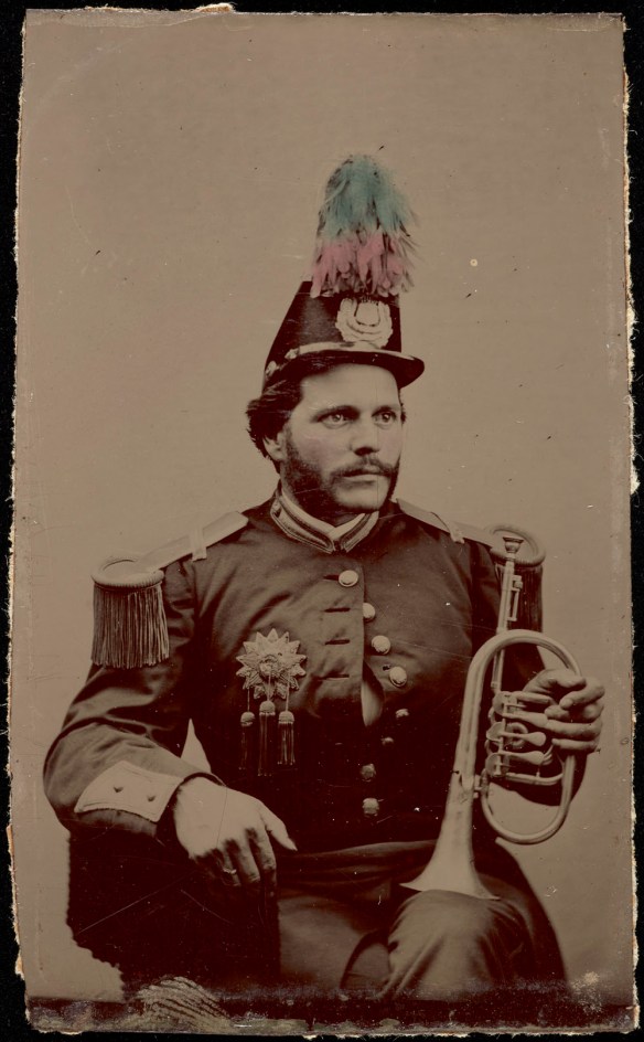 Photo noir et blanc d’un homme assis, vêtu d’un uniforme d’harmonie et d’un chapeau orné de plumes. L’homme tient un bugle (clairon) contre son genou gauche.